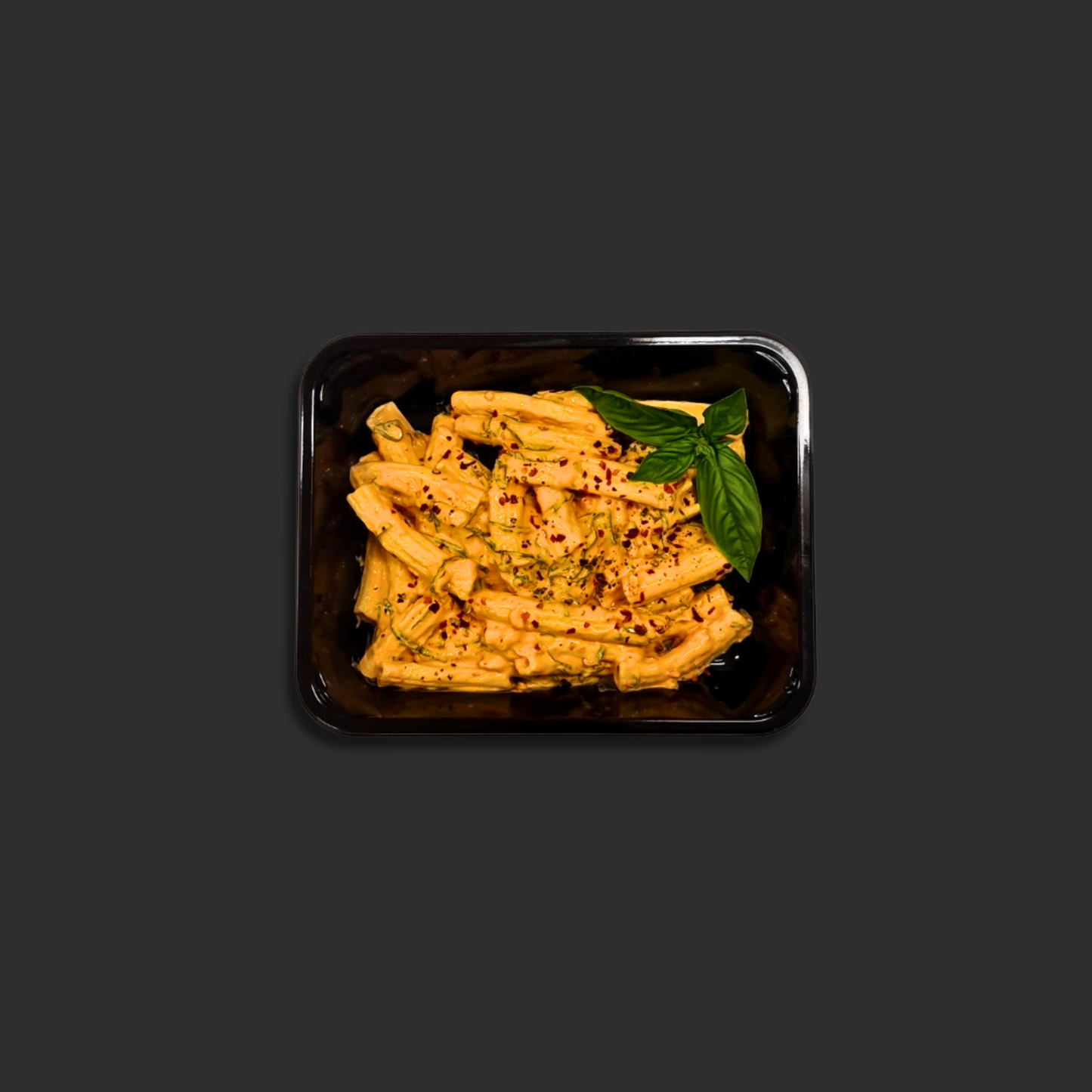 Vodka Rigatoni served in a black ready meal container with pasta in creamy tomato vodka sauce, chili flakes and basil garnish in the corner.